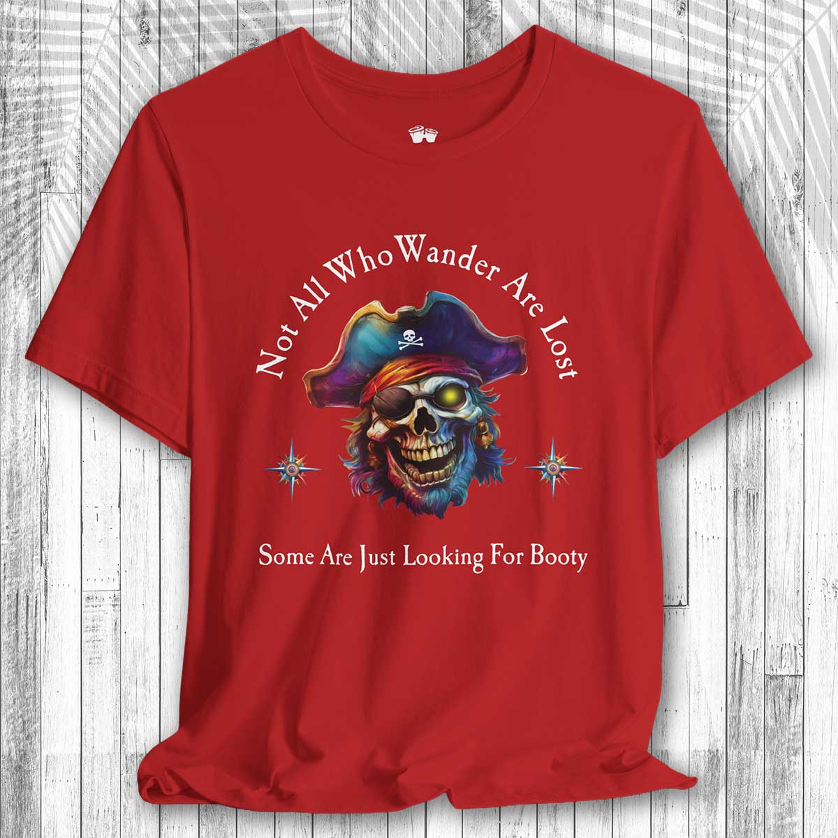 Red t-shirt with a pirate-themed design and the text: "Not All Who Wander Are Lost – Some Are Just Looking For Booty"  on a wooden surface