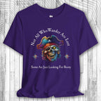 Purple t-shirt with a pirate skull design and the text: "Not All Who Wander Are Lost – Some Are Just Looking For Booty" on a wooden surface