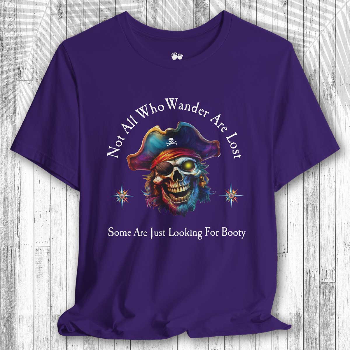 Purple t-shirt with a pirate skull design and the text: "Not All Who Wander Are Lost – Some Are Just Looking For Booty" on a wooden surface