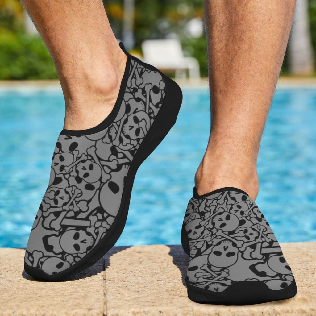 A person wearing gray and black slip-on water shoes with a skull and crossbones pattern, standing on a pool deck with a swimming pool in the background.