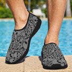 A person wearing gray and black slip-on water shoes with a skull and crossbones pattern, standing on a pool deck with a swimming pool in the background.