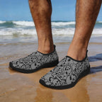 Gray slip-on water shoes with a black and white pirate's skull and crossbones pattern, worn by a person standing on a wet sandy beach with the ocean in the background.