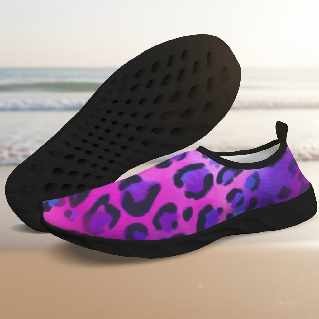A pair of slip-on water shoes with a vibrant pink and purple leopard print upper and black soles, displayed against a blurred beach background.