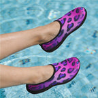 A pair of slip-on water shoes with a purple and pink leopard print pattern and black soles, shown being worn on feet in a swimming pool.