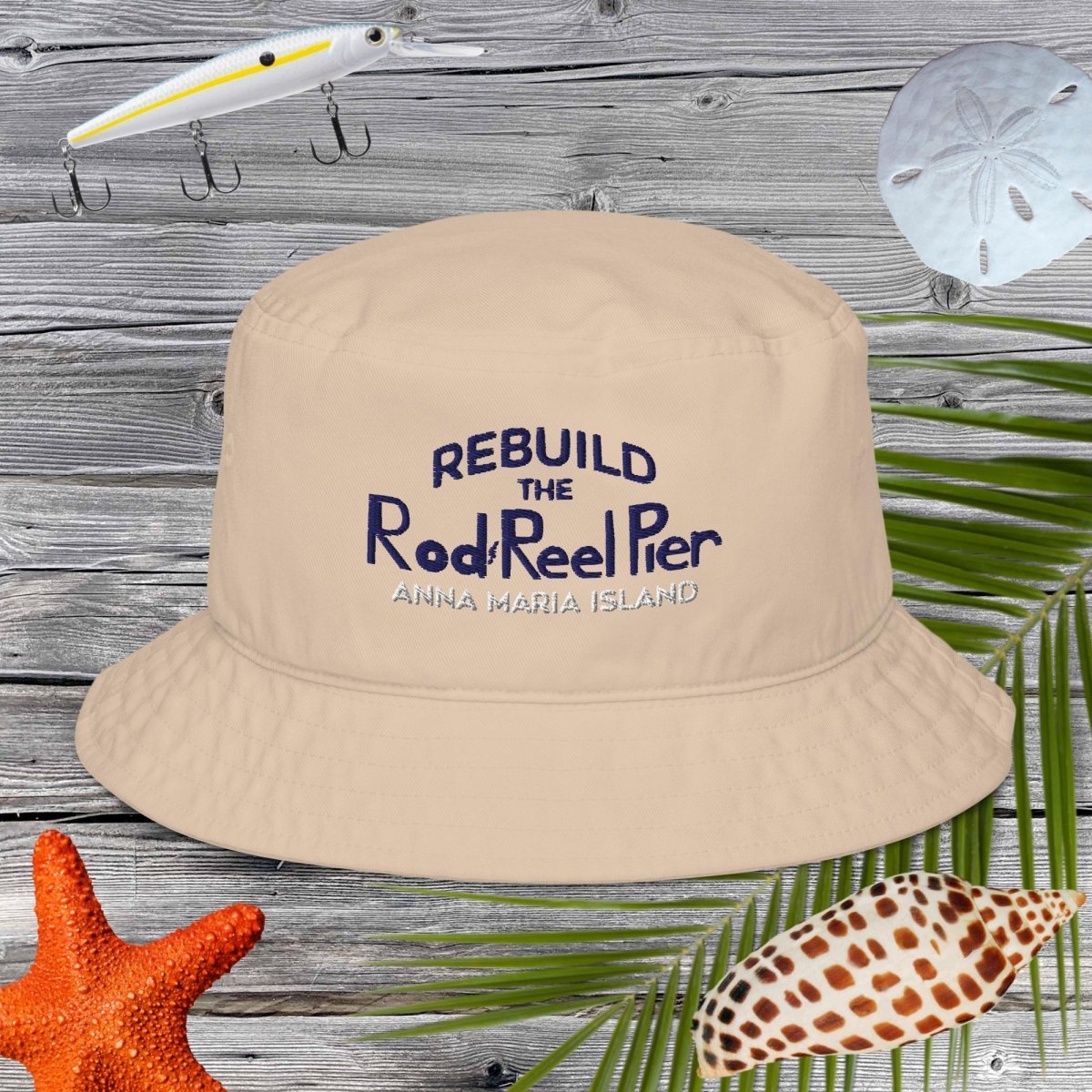 Beige bucket hat with Rebuild the Rod & Reel Pier, Anna Maria Island text on a wooden surface with fishing-related items.
