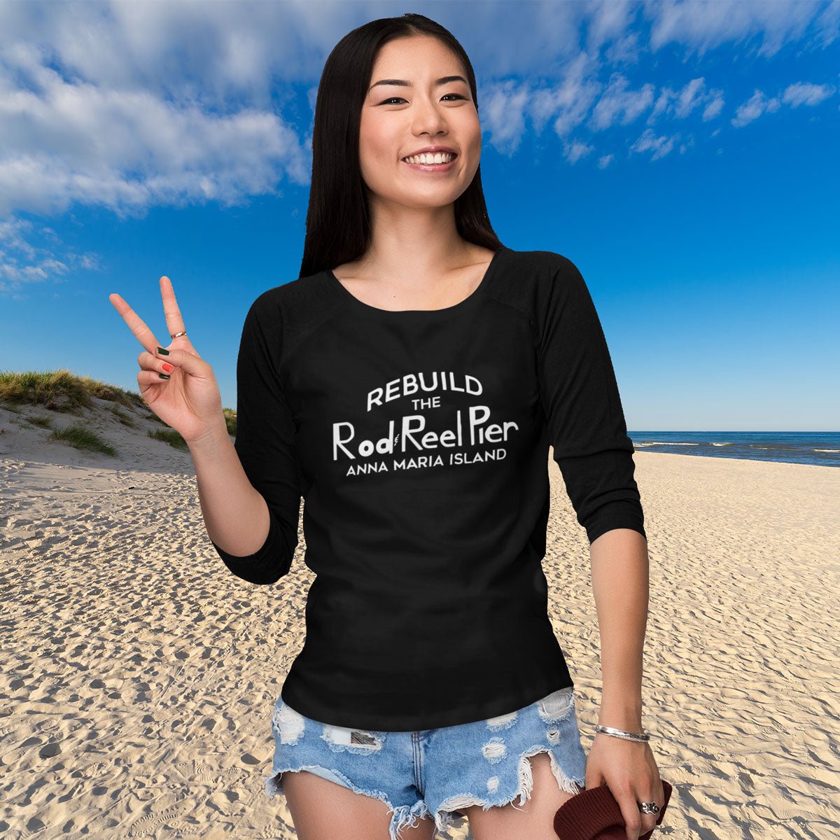 Woman on a beach wearing a black t-shirt with text, making a peace sign.