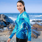 Woman wearing a blue wipeout patterned rashie standing on a beach with ocean and rocks in the background