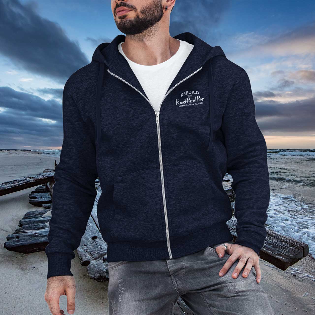 Man wearing a navy blue hoodie with a brand logo, standing on a dock with a cloudy sky and water background.