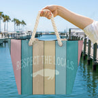 Colorful tote bag with 'Respect the Locals' text and Manatee graphic held out by a woman's hand with a West Florida Marina in the background.