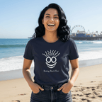 A woman wearing a navy blue t-shirt with a white graphic of a funny face and the text 'Resting Beach Face', standing against a rustic wooden wall.