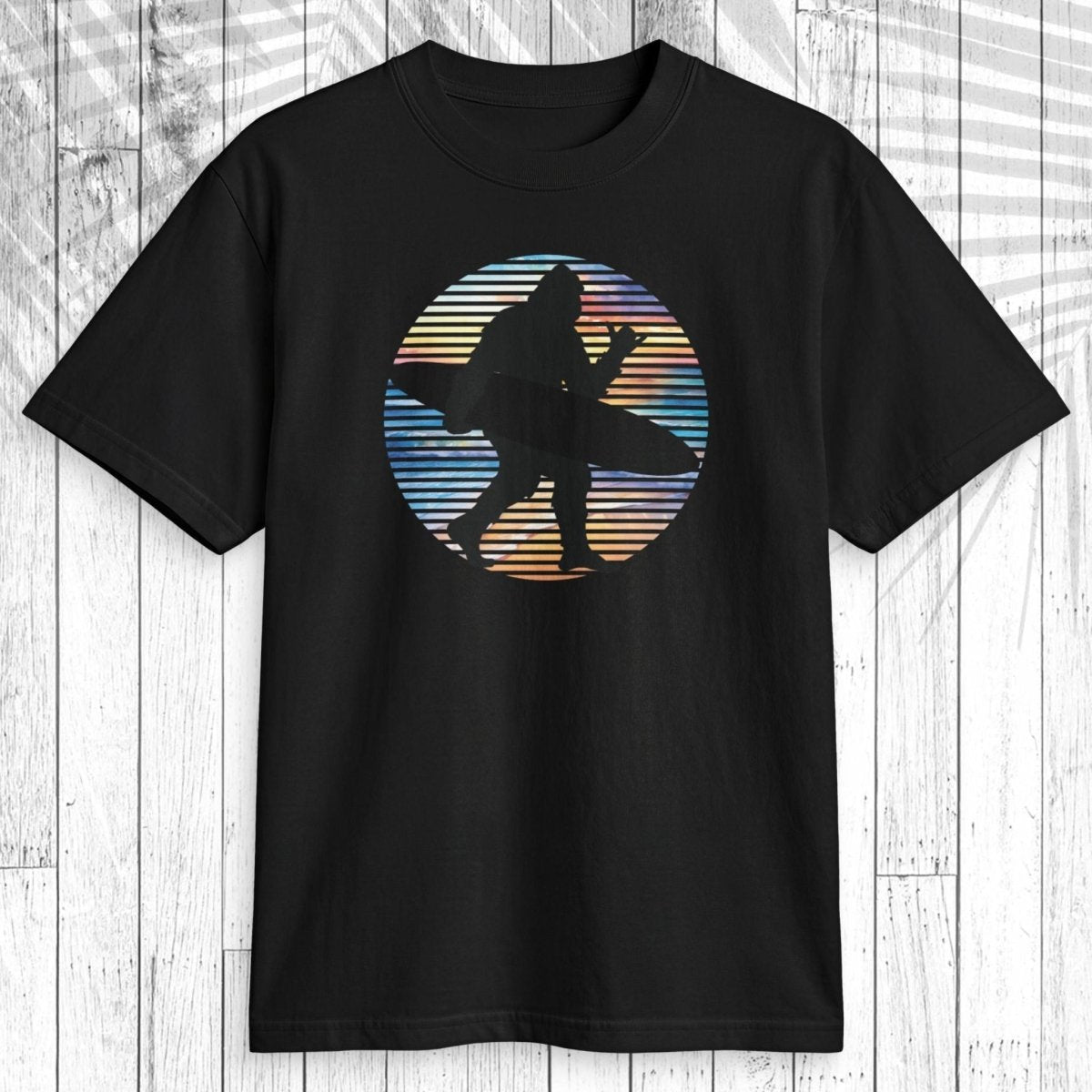 A black short-sleeve crewneck t-shirt featuring a circular graphic of a sasquatch surfer silhouette against a retro striped sunset background, displayed flat on a white wood plank surface.