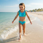 Happy little girl wearing a Two-piece Sea Turtle Bikini with cute turtle print running along the shore line splashing in the waves