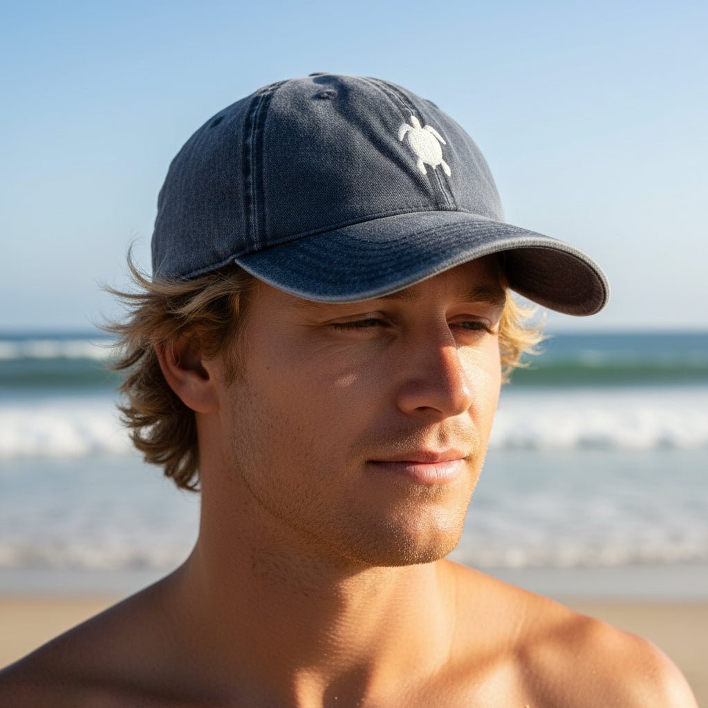 A man on a beach wearing a navy blue baseball cap with a white sea turtle logo on the front, with the ocean and sky blurred in the background.