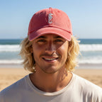 A smiling man with blonde hair wearing a coral pink baseball cap with a white seahorse logo, standing on a sandy beach with the ocean in the background.