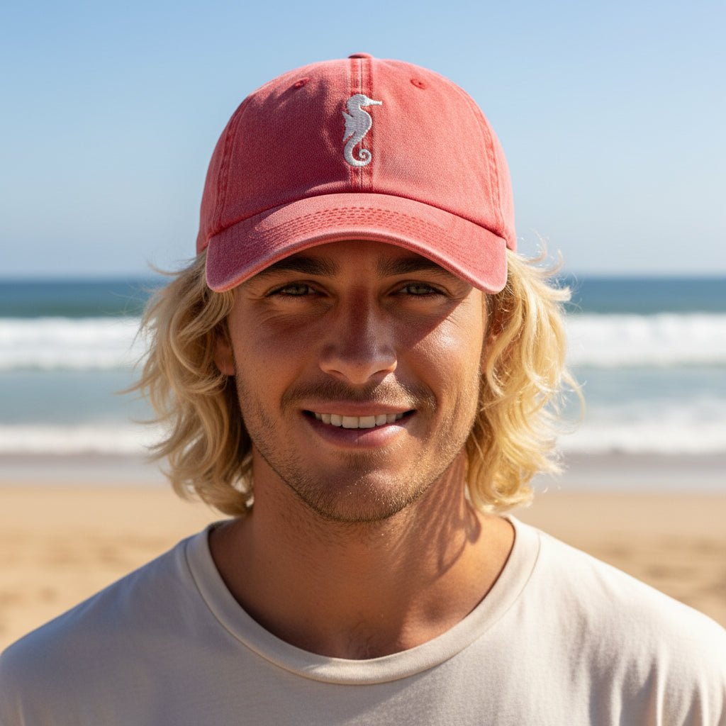 A smiling man with blonde hair wearing a coral pink baseball cap with a white seahorse logo, standing on a sandy beach with the ocean in the background.