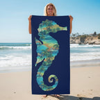 A rectangular beach towel with a dark navy blue background, featuring a large seahorse silhouette filled with a tropical palm tree and wave pattern in shades of teal, green, and yellow.