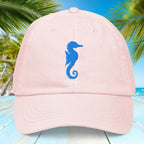Pink pastel cap with a blue seahorse embroidery on a tropical background with palm trees and blue water.