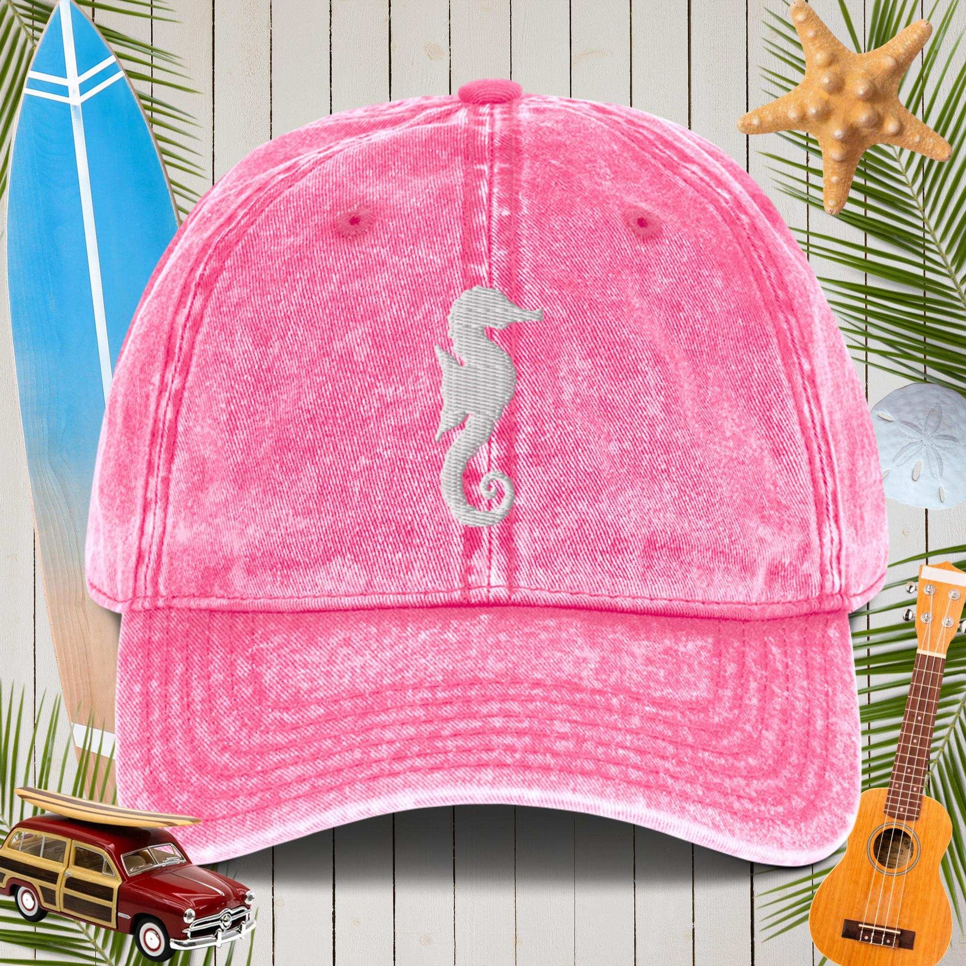 Pink cap with a seahorse design on a beach-themed background with surfboard, car, and ukulele.