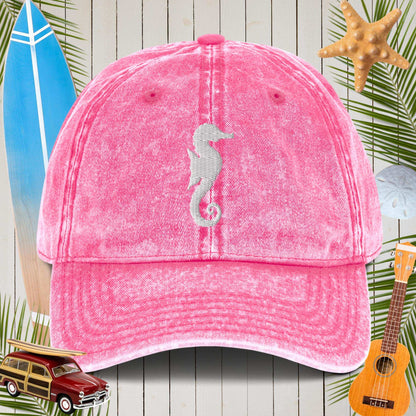 Pink cap with a seahorse design on a beach-themed background with surfboard, car, and ukulele.