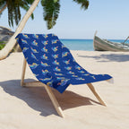 A blue beach towel with a repeating pattern of colorful shaka signs is draped over a wooden lounge chair on a sandy beach with palm trees and the ocean in the background.