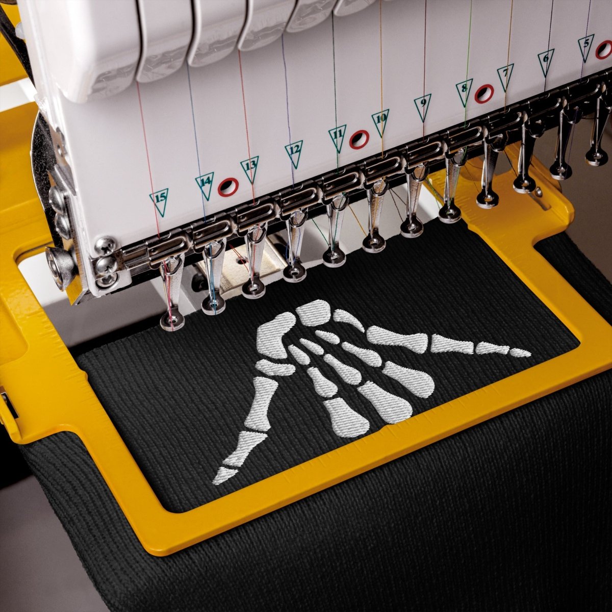 Close-up of an embroidery machine stitching a white shaka sign skeleton hand design onto black fabric, with the yellow machine frame and embroidery needles clearly visible.
