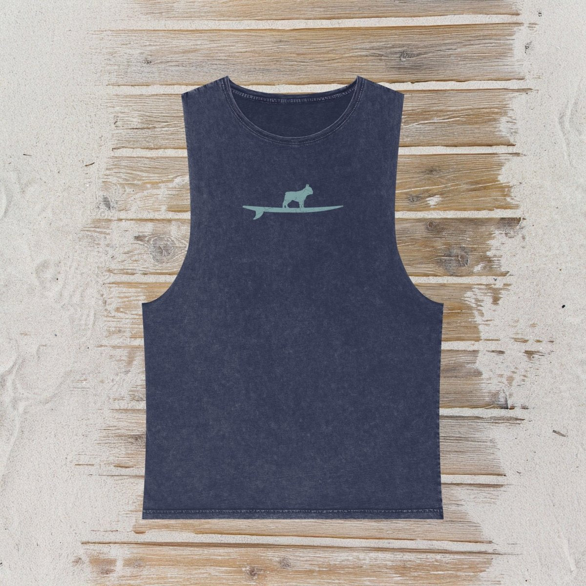 A sleeveless tank top in a washed navy blue color, featuring a small light gray graphic of a frenchy standing on a surfboard on the chest. The tank top is laid flat on a rustic wooden plank surface.