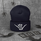 A dark navy blue knit beanie with a white skeleton hand graphic on the cuff, displayed on a textured gray background with Halloween-themed props including spiderwebs, bat wings, and 'BOO' letter cutouts.
