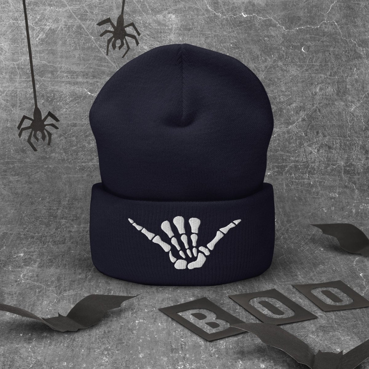 A dark navy blue knit beanie with a white skeleton hand graphic on the cuff, displayed on a textured gray background with Halloween-themed props including spiderwebs, bat wings, and 'BOO' letter cutouts.