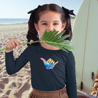 Young girl on a beach holding a leaf, wearing a black Long Sleeve Toddler Tee with a colorful Shaka design.