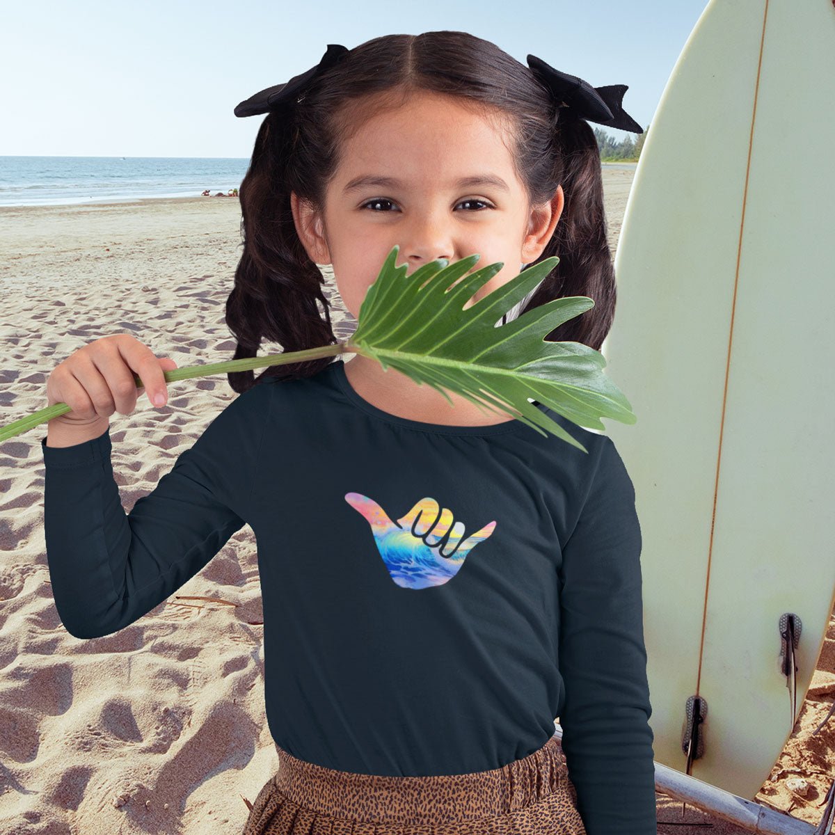 Young girl on a beach holding a leaf, wearing a black Long Sleeve Toddler Tee with a colorful Shaka design.