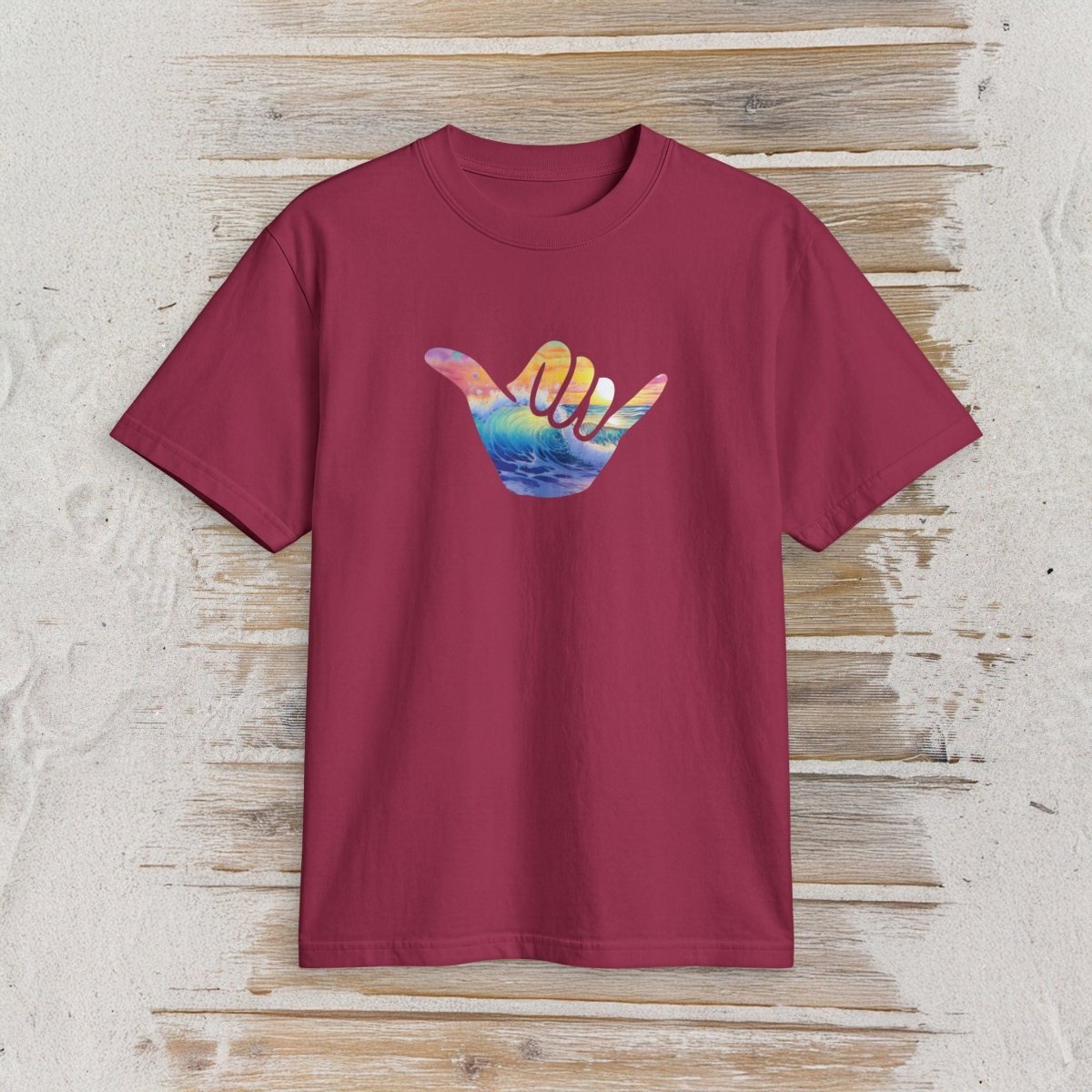 A short-sleeve crewneck t-shirt in maroon, featuring a colorful graphic of a hand sign with a sunset and ocean design, displayed flat against a rustic light wood background.