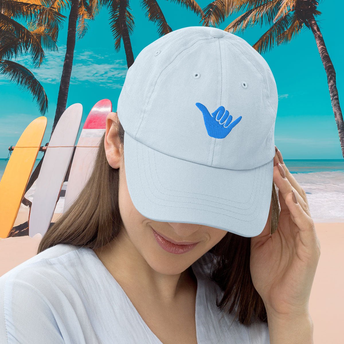 Person wearing a light Pastel Baseball Hat with a blue Shaka logo against a beach background with surfboards and palm trees.