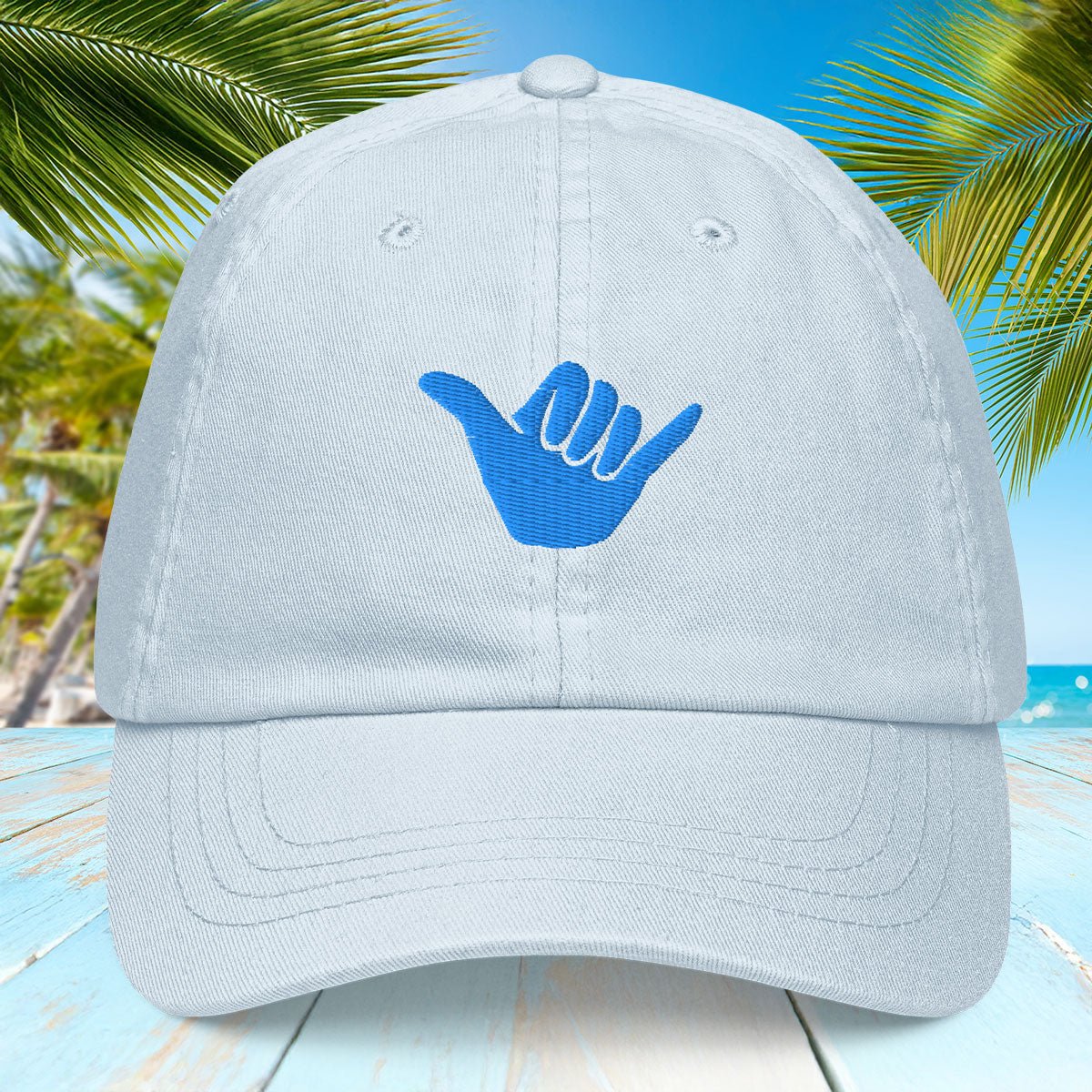 Light pastel blue cap with blue 'shaka' hand symbol on a tropical background