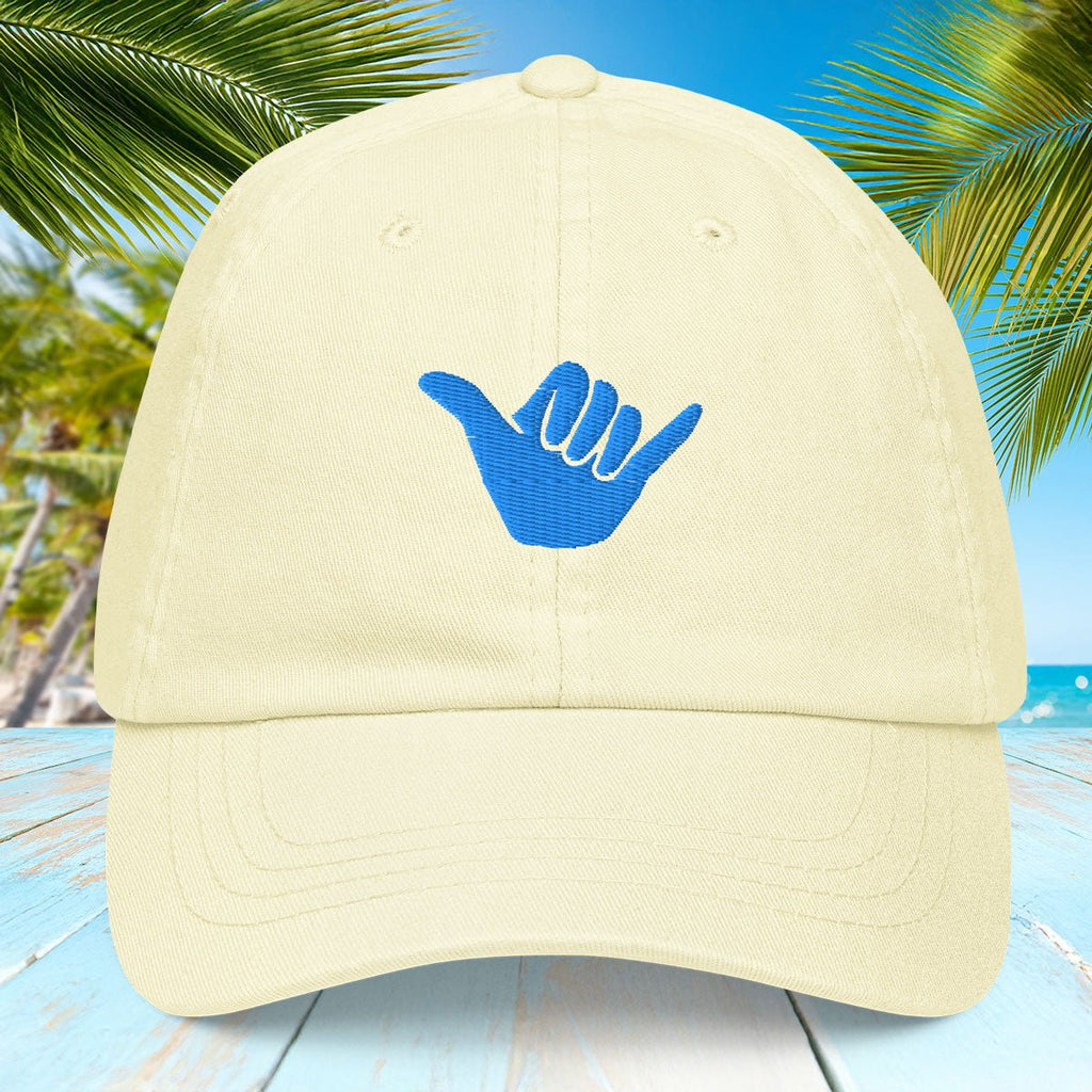 Pastel lemon yellow cap with blue Shaka symbol on a tropical background