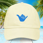 Pastel lemon yellow cap with blue Shaka symbol on a tropical background