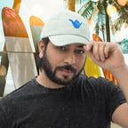 Man wearing a pastel mint green cap with a blue Shaka logo, standing in front of colorful surfboards and palm trees.