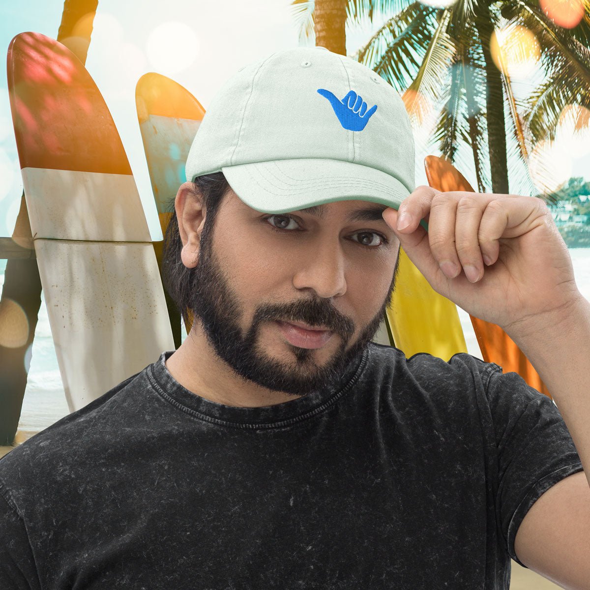 Man wearing a pastel mint green cap with a blue Shaka logo, standing in front of colorful surfboards and palm trees.