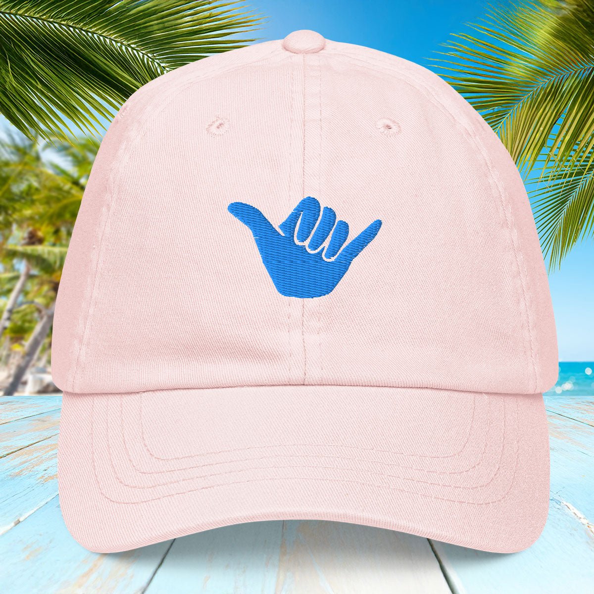 Pastel Pink cap with blue Shaka symbol against a tropical background