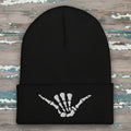 A black ribbed knit beanie hat with a folded cuff featuring a white embroidered skeleton hand graphic in a shaka sign pose, shown on a rustic wood background