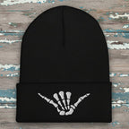 A black ribbed knit beanie hat with a folded cuff featuring a white embroidered skeleton hand graphic in a shaka sign pose, shown on a rustic wood background