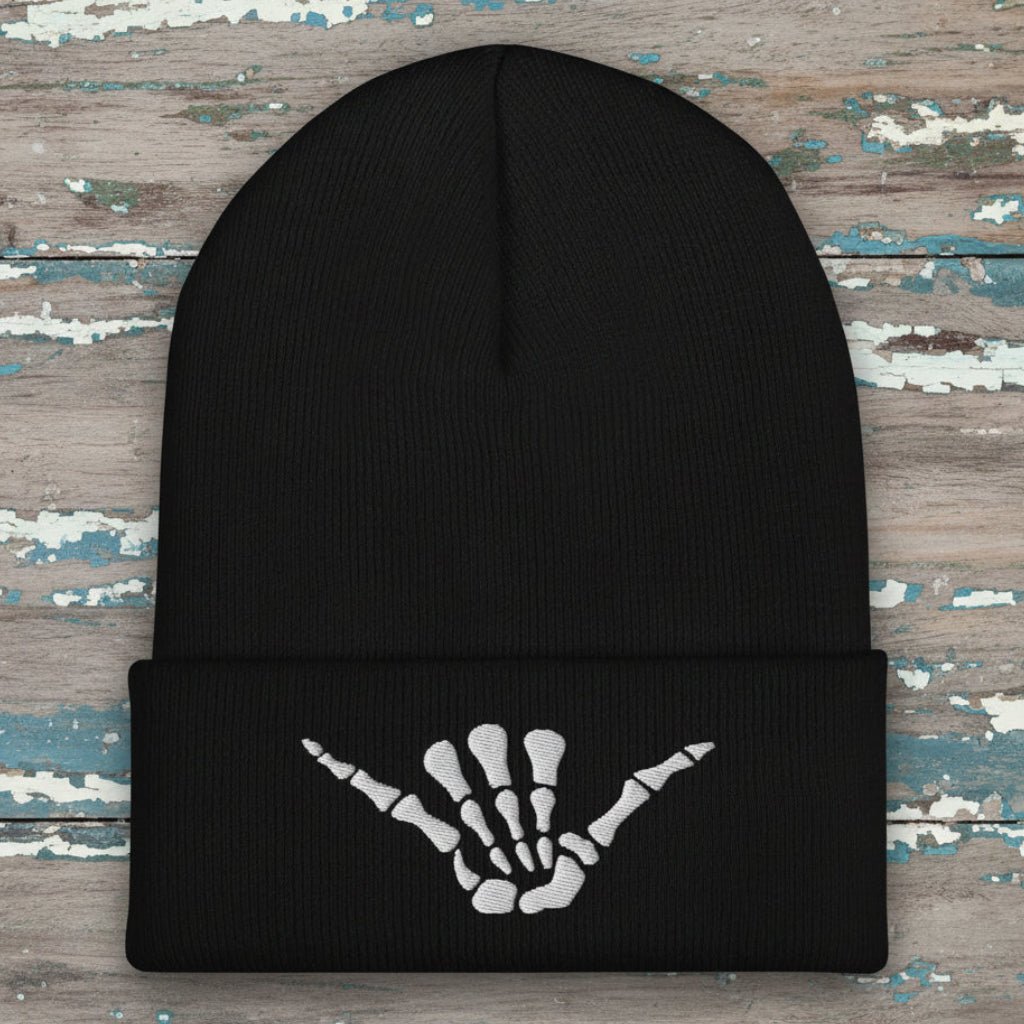 A black ribbed knit beanie hat with a folded cuff featuring a white embroidered skeleton hand graphic in a shaka sign pose, shown on a rustic wood background