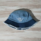 Blue bucket hat with frayed edge and embroidered shaka bones on a rustic wood surface
