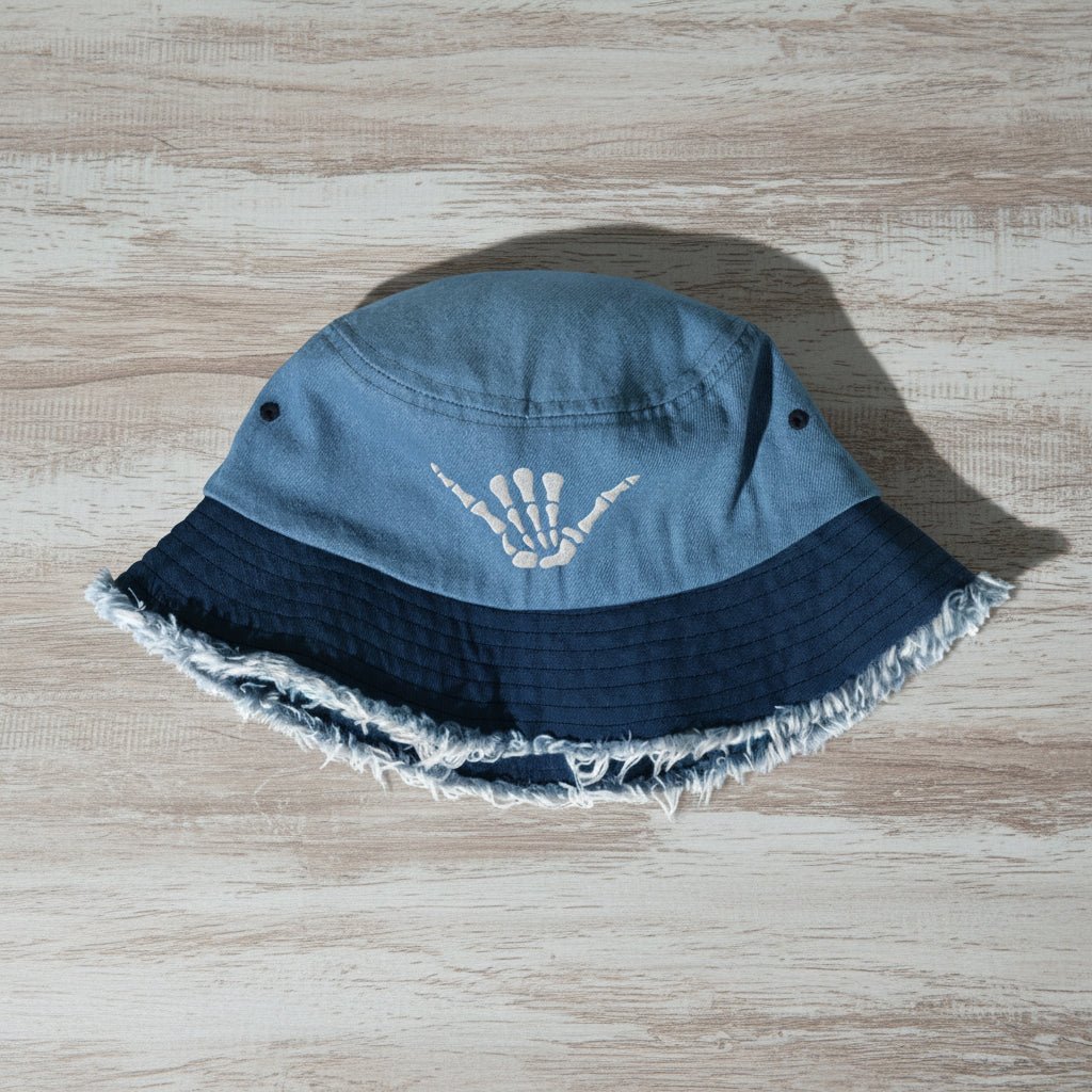 Blue bucket hat with frayed edge and embroidered shaka bones on a rustic wood surface
