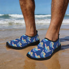 A person standing on a sandy beach wearing slip-on water shoes with a blue background and a repeating multicolor Shaka graphic pattern. The ocean waves and blue sky are visible in the background.