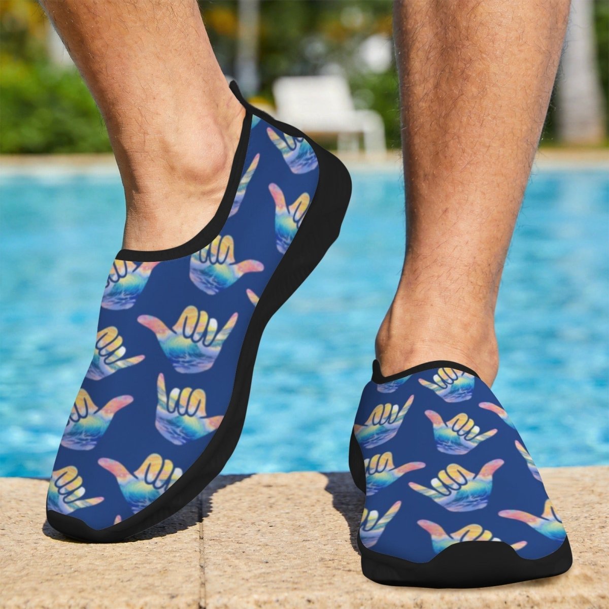 A person standing by a swimming pool wearing Shaka Surf slip-on water shoes with a blue background and a repeating multicolor Shaka hand graphic pattern.