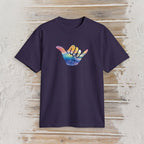 A dark purple short-sleeve t-shirt with a colorful Hawaiian 'shaka' hand graphic on the chest, displayed flat against a rustic light wood plank background.