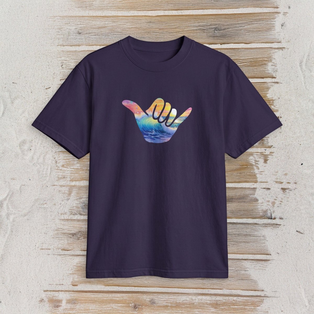 A dark purple short-sleeve t-shirt with a colorful Hawaiian 'shaka' hand graphic on the chest, displayed flat against a rustic light wood plank background.