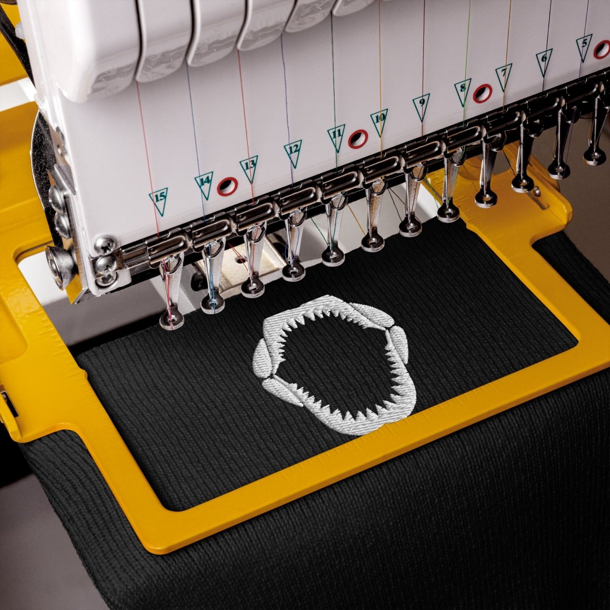 Close-up of an embroidery machine stitching a white geometric patch onto black fabric, with the yellow and white machine body visible.