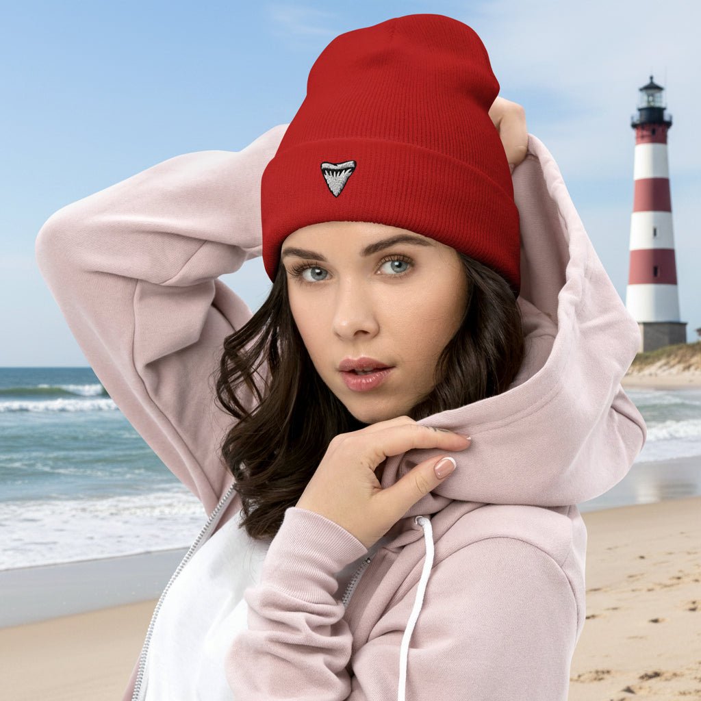 Woman wearing a red beanie with a shark tooth embroidered logo and a pink hoodie on a beach near a lighthouse