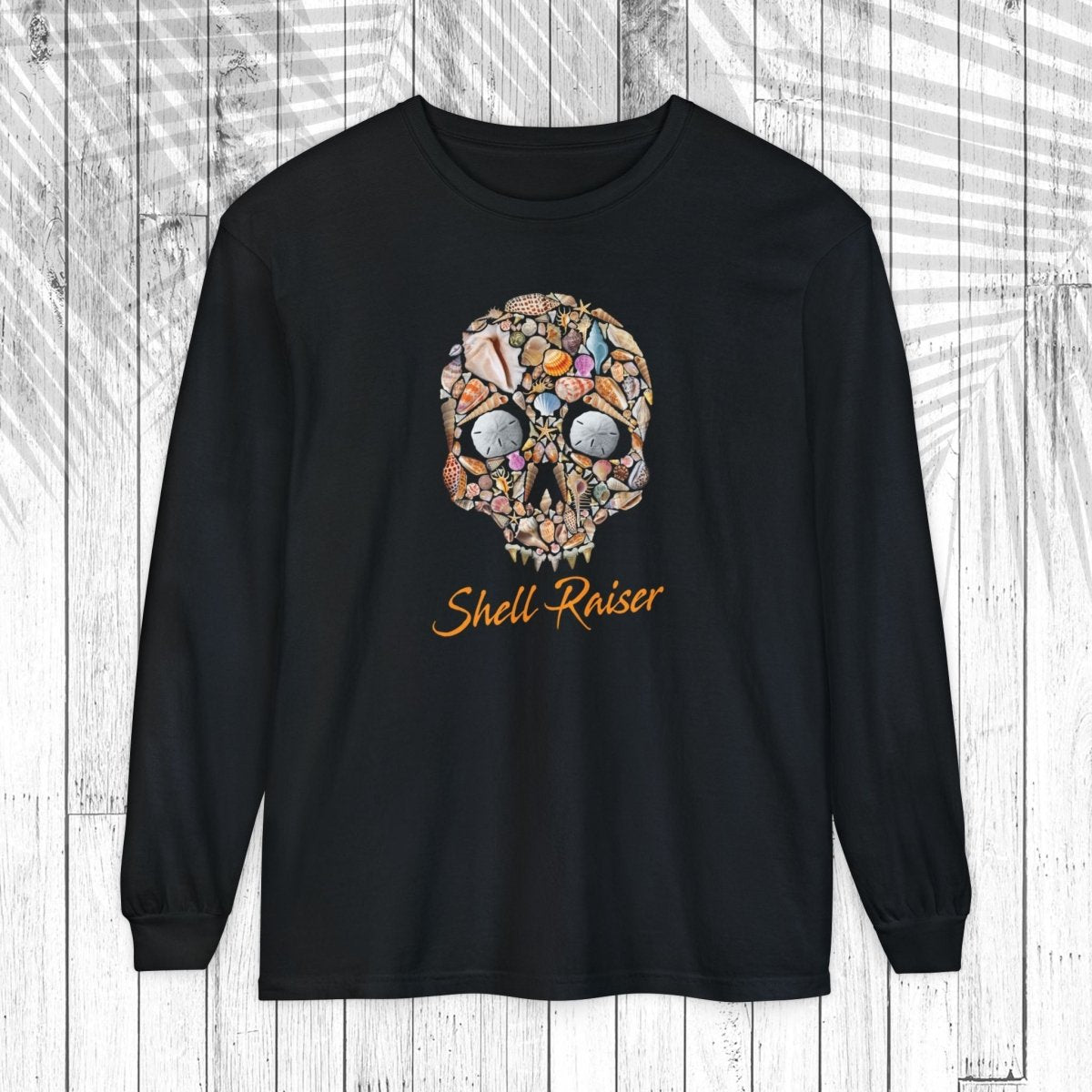 A black long-sleeve t-shirt with a colorful skull graphic made of various small objects and the text 'Shell-Raiser' in orange cursive, displayed on a white wood plank background.